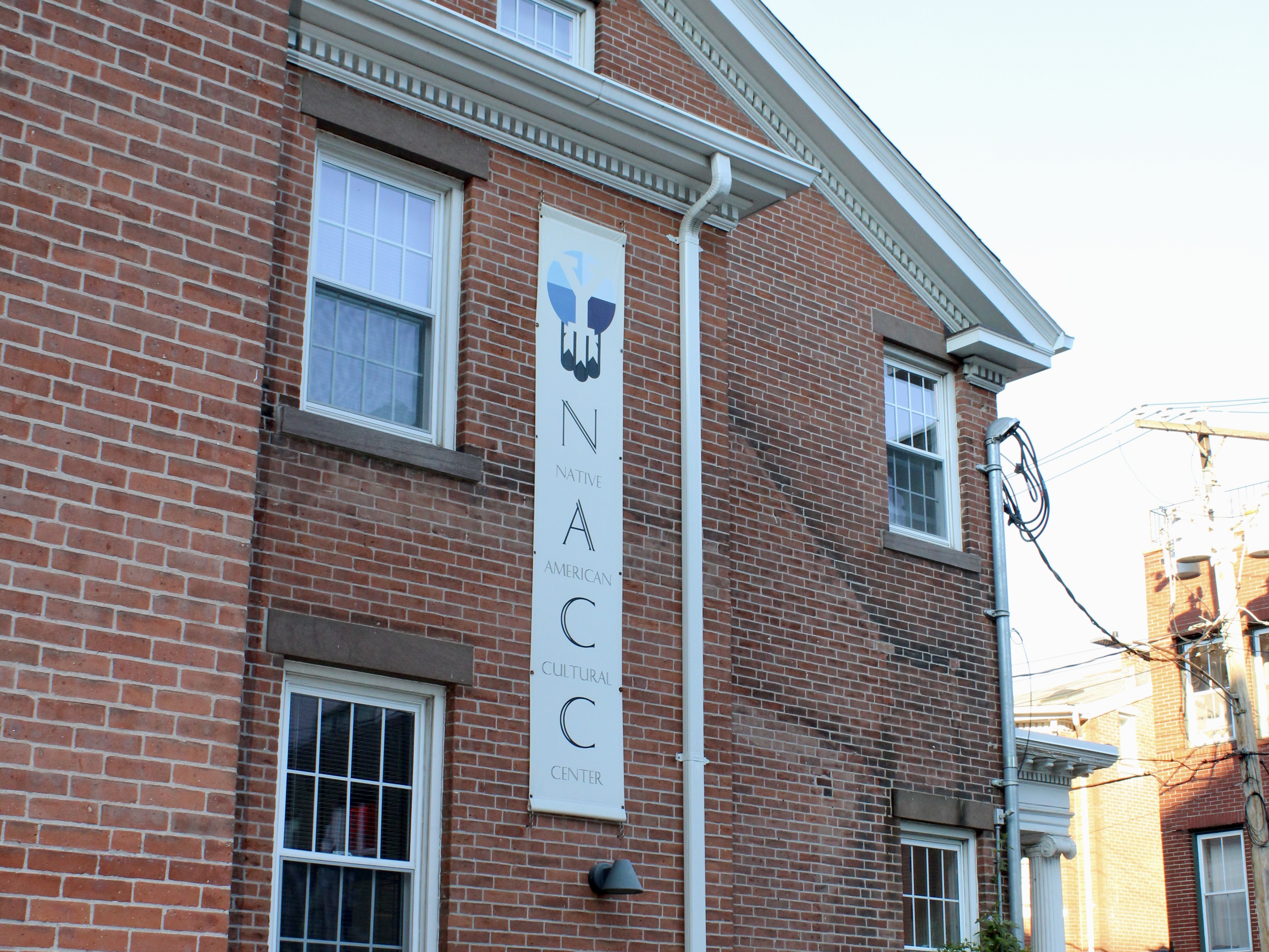 Exterior view of the Native American Cultural Center (NACC) at Yale, a red-brick, three-story building with a vertical NACC banner displayed near the entrance